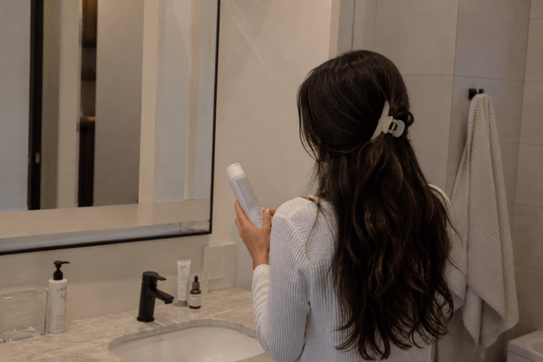 Woman with long dark hair at a bathroom mirror holding a hair product, the residue that migrates from hair to pillowcase to forehead overnight