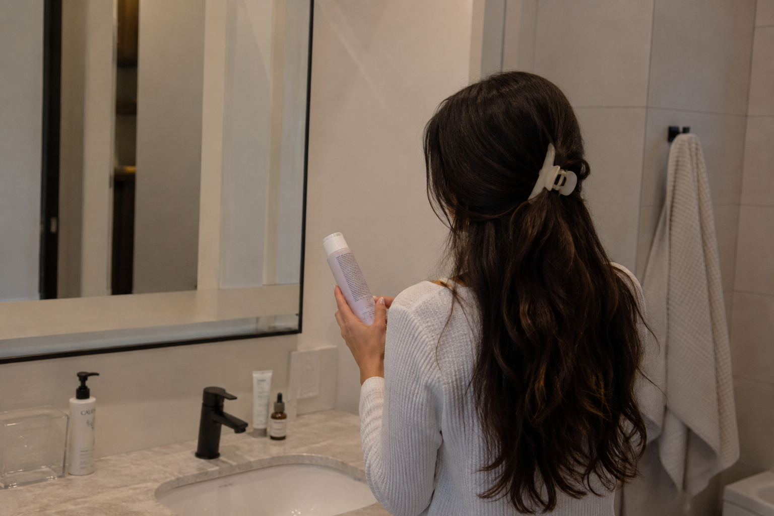 A woman with long dark brown hair is seen from behind, holding a white bottle in a bathroom setting with a sink, mirror, and towel visible.
